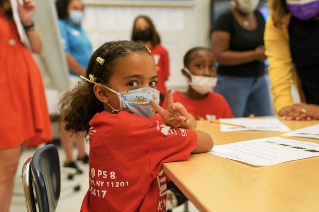 A young girl wearing a red tshirt and a face mask gives the peace sign to the camera at PS 8 in Brooklyn.